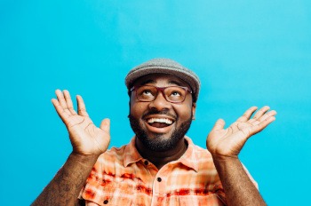 Man in orange checkered shirt and gray hat lifting hands and smiling with light blue background